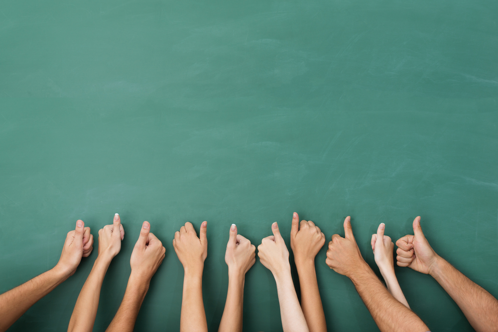 Close up view of the hands of a group of people giving a thumbs up gesture of approval an success with their hands raised against a blank green chalkboard with copyspace