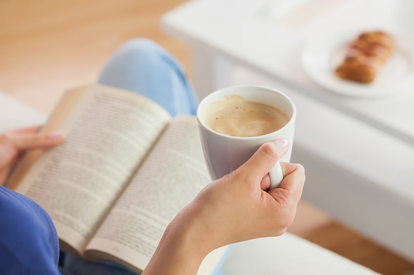 Woman sitting on the sofa reading a book holding her coffee mug in living room at home.